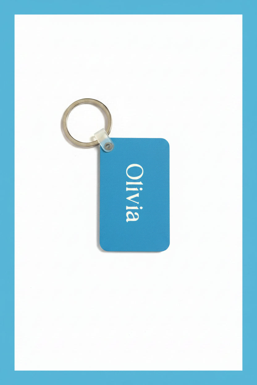 A rectangular blue keychain tag with rounded corners, featuring the name 'Olivia' printed vertically in white. The tag is attached to a silver metal split keychain against a plain white background with a blue border. Roxzell