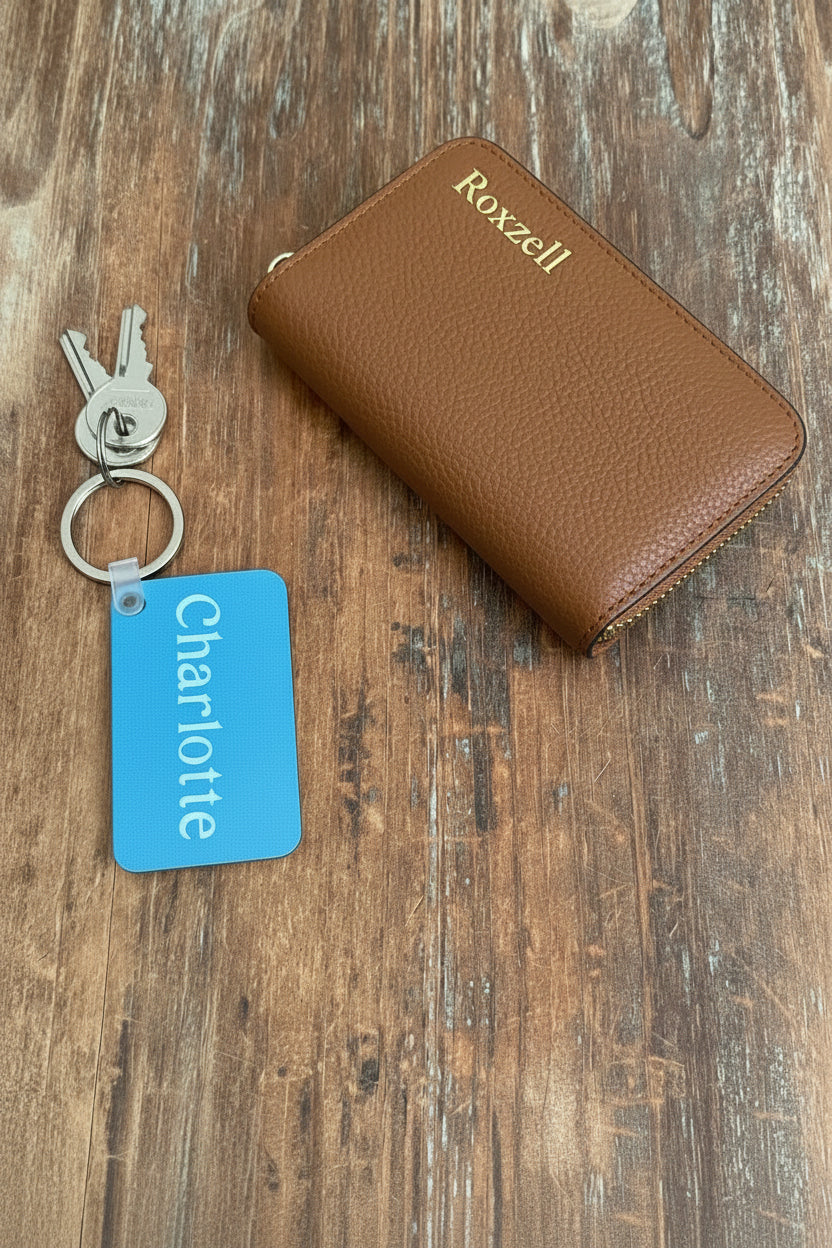 A light blue keychain with name 'Charlotte' beside a tan leather key case placed on a rustic wooden surface. Roxzell