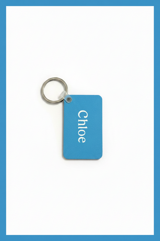 A personalized light blue rectangular key tag with rounded corners, featuring the name 'Chloé' printed vertically in white, attached to a silver metal split key ring, set against a white background with a blue border. Roxzell