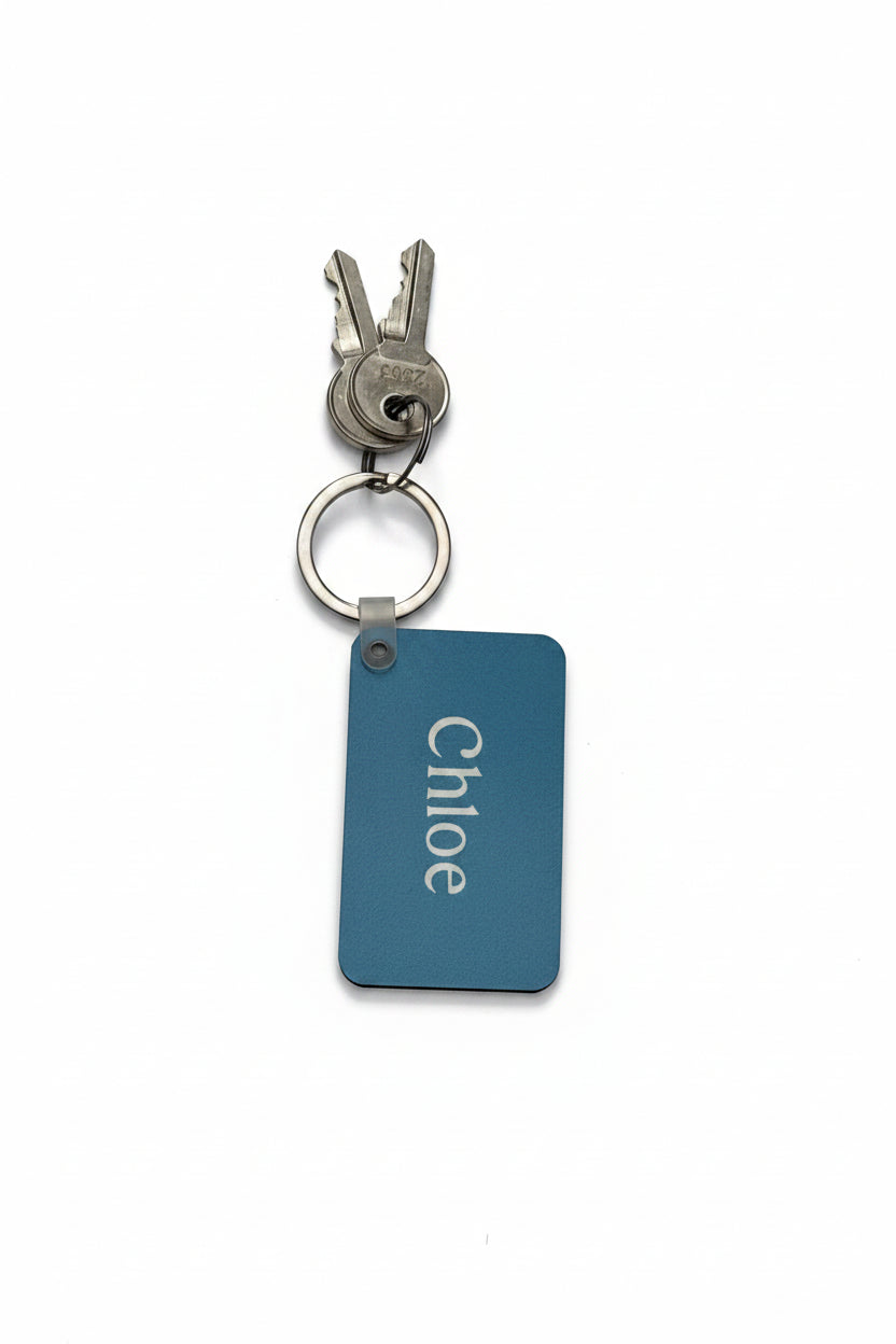 A personalized keychain with a rectangular blue keychain with the name 'Chloe' in white, attached to a silver split ring and keys, on a white background. Roxzell