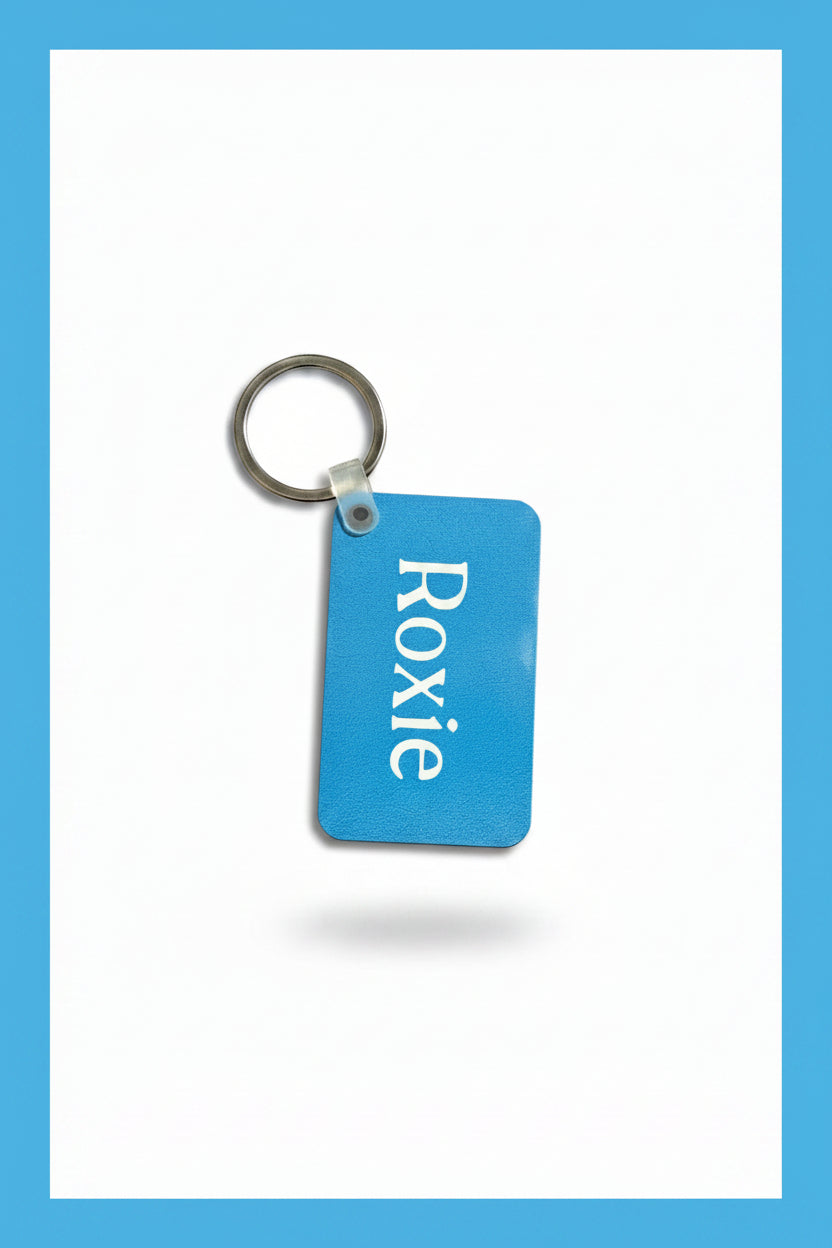 A light blue rectangular keychain with the name 'Roxie' printed in white, attached to a silver metal key ring, placed on a white background with a light blue border. Roxzell