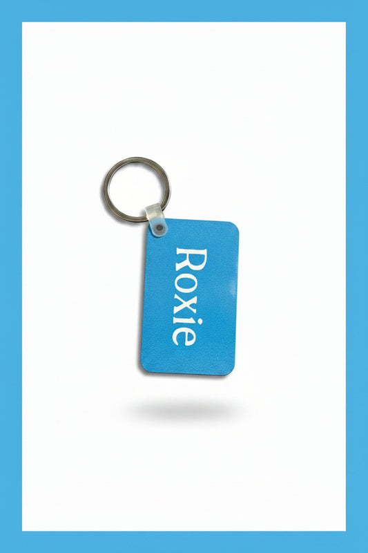 A light blue rectangular keychain with the name 'Roxie' printed in white, attached to a silver metal key ring, placed on a white background with a light blue border. Roxzell