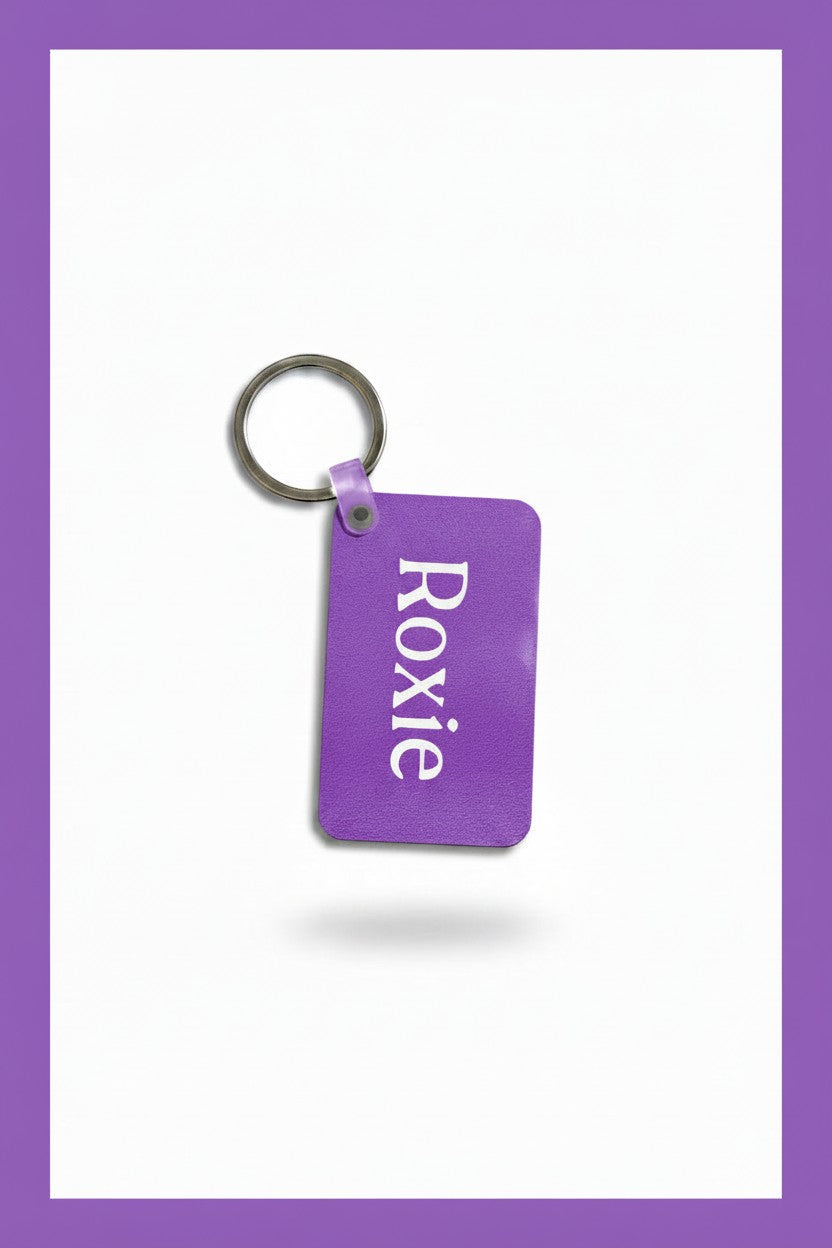 A purple rectangular keychain tag with rounded corners, featuring the name 'Roxie' printed vertically in white. The tag is attached to a silver metal split ring and is shown against a white background with a purple border. Roxzell