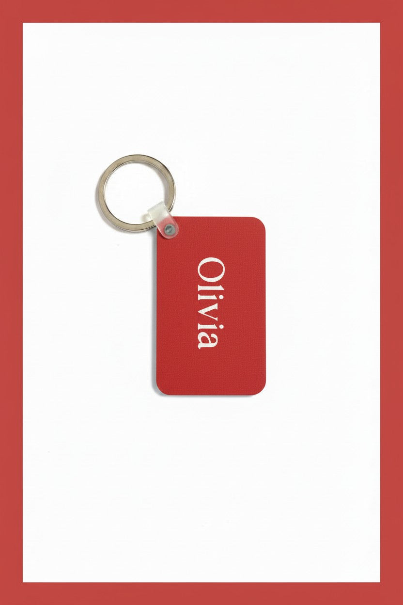 A red rectangular keychain tag with the name 'Olivia' printed vertically in white, attached to a silver metal key ring, set against a white background with a red border. Roxzell