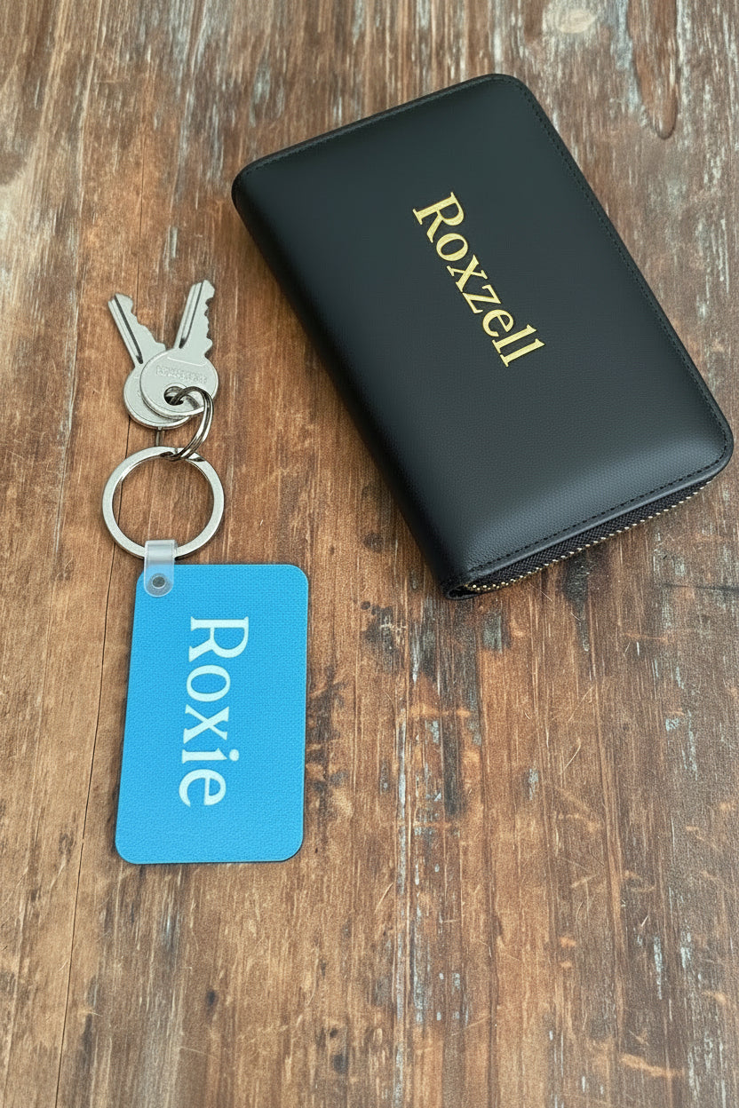 A blue keychain with name 'Roxie' and two keys beside a black leather Roxzell wallet, shown on a rustic wooden background. Roxzell 