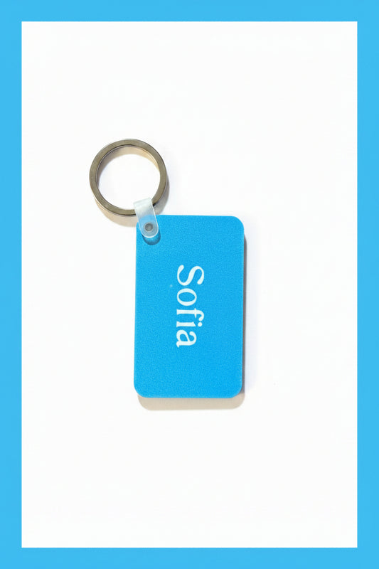 A light blue rectangular keychain with the name 'Sofia' printed in white, attached to a silver metal key ring, placed on a white surface. Roxzell