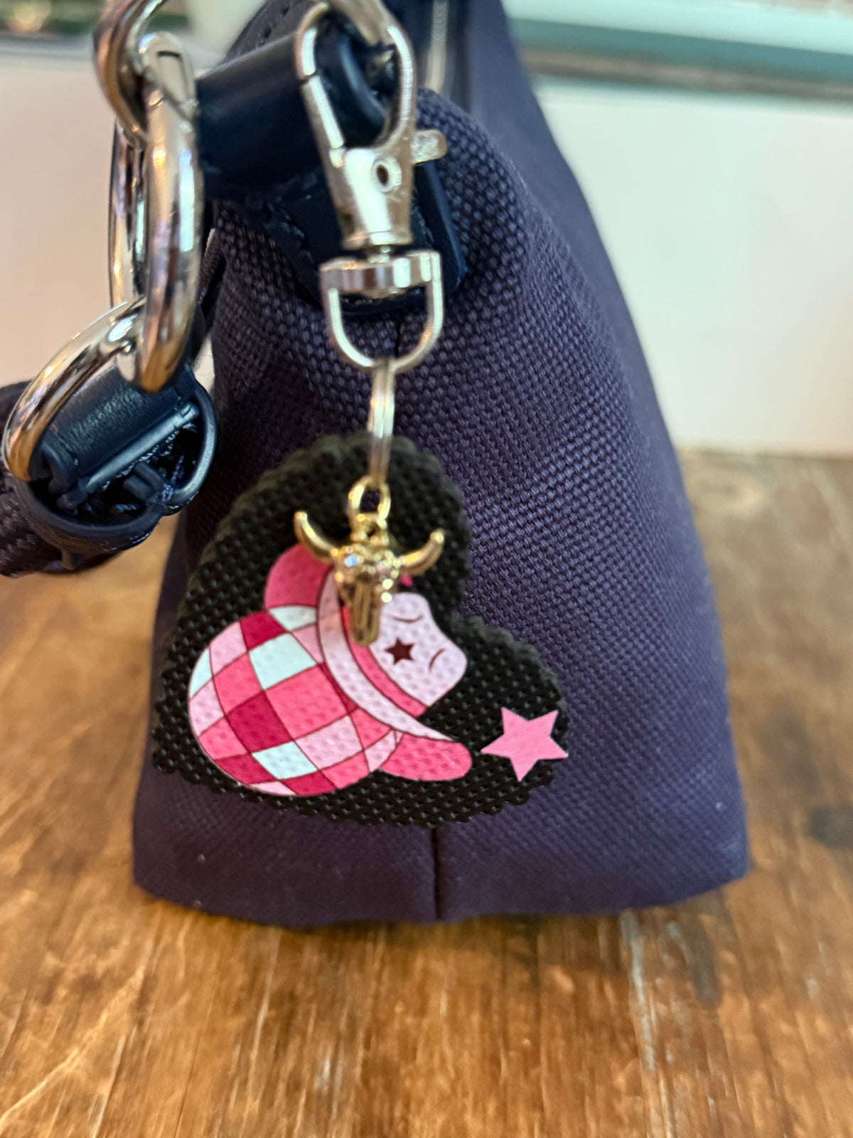 A close-up of bag charm with a pink and white checkered cowboy hat, a gold-toned bull charm, and a pink star, hanging from silver metal rings on a navy fabric handbag on a wooden surface. Roxzell
