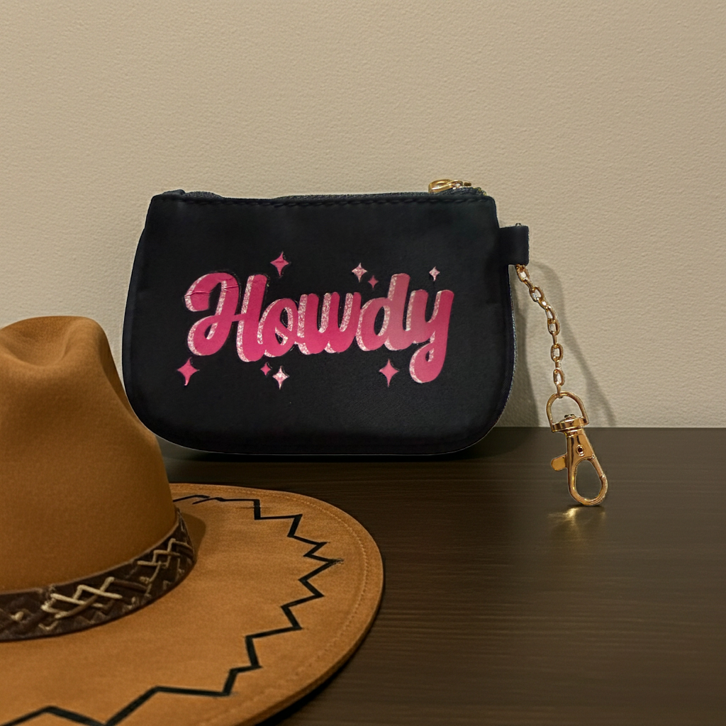 Black coin purse/id wallet with 'Howdy' text and keychain on a wooden surface with a brown cowboy hat.