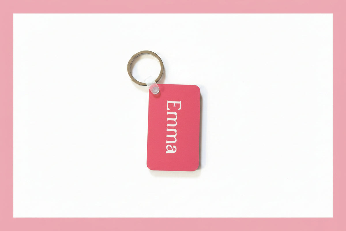 A pink rectangular keychain with the name 'Emma' in white script, attached to a silver metal key ring, placed on a rustic wooden surface.