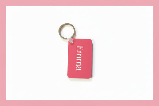 A pink rectangular keychain with the name 'Emma' in white script, attached to a silver metal key ring, placed on a rustic wooden surface.