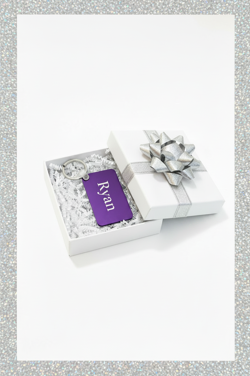 A personalized purple keychain with the name 'Ryan' in white script, presented inside an open white gift box with white crinkle paper and a silver ribbon bow on a white background and silver border. Roxzell