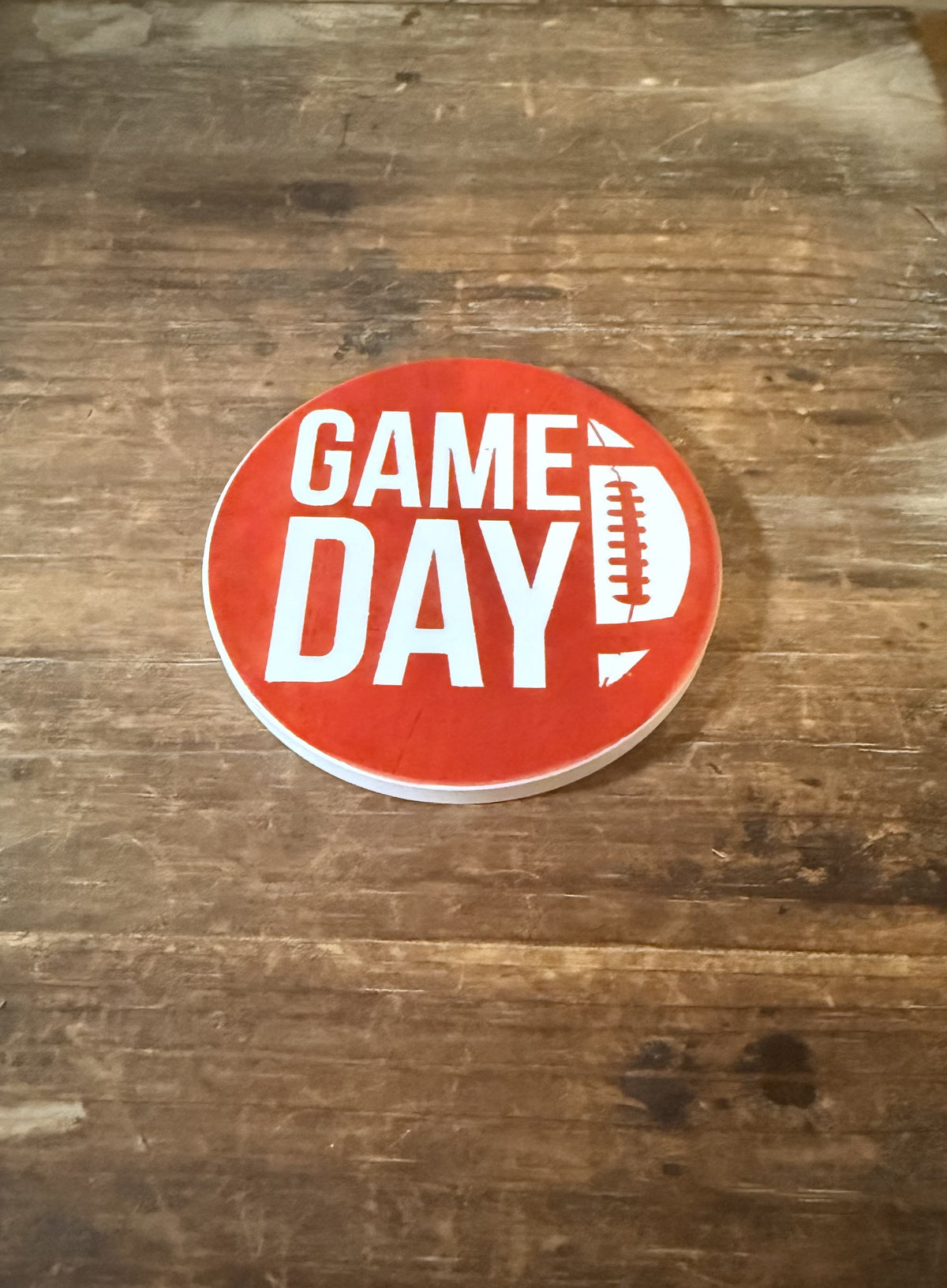 Round red coaster with bold white “GAME DAY” text and football‑shaped letter D on rustic wood background. Roxzell