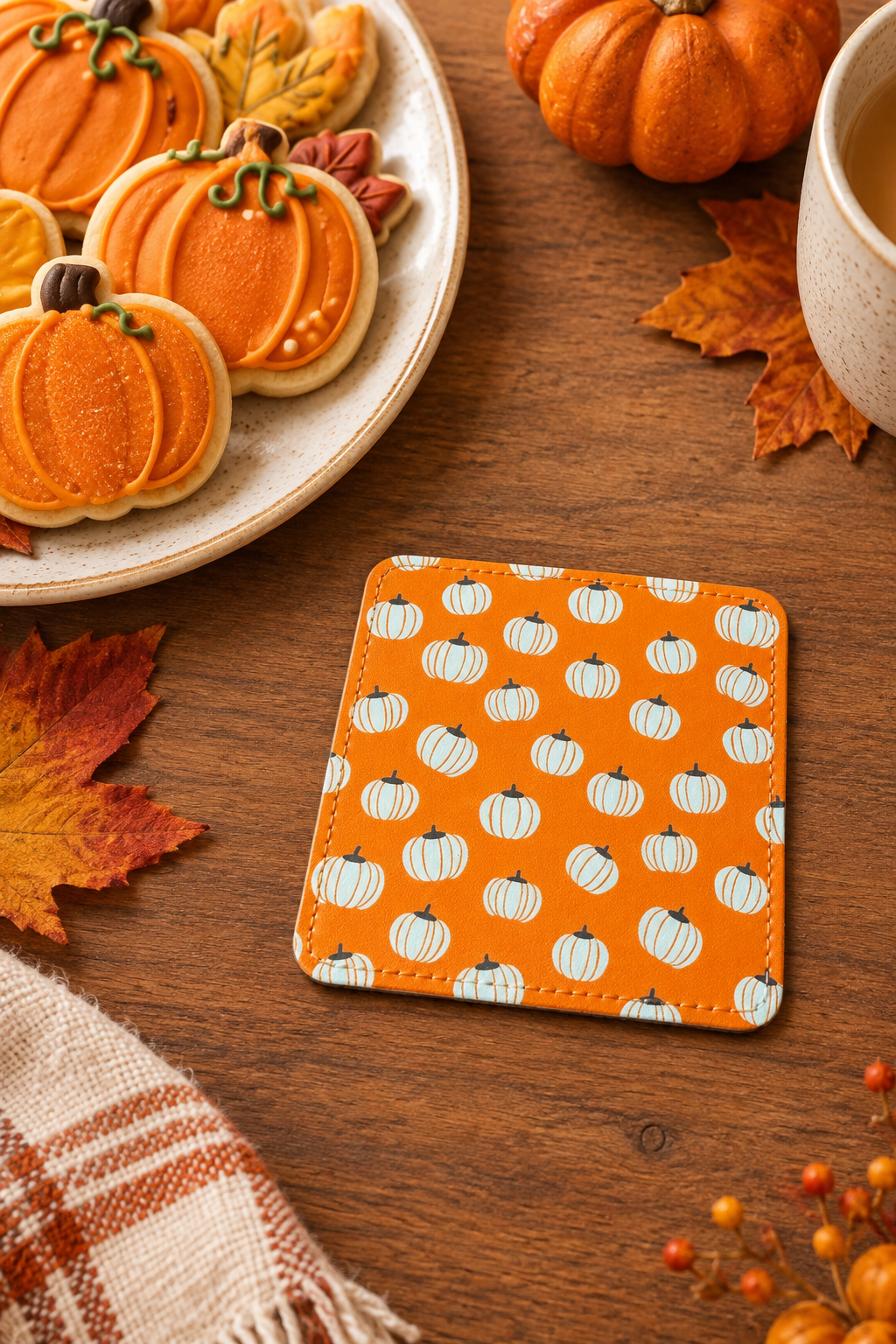 An orange square coaster with a white pumpkin pattern, styled on a wooden table next to autumn leaves, a plate of pumpkin cookies, and a cup of coffee. Roxzell