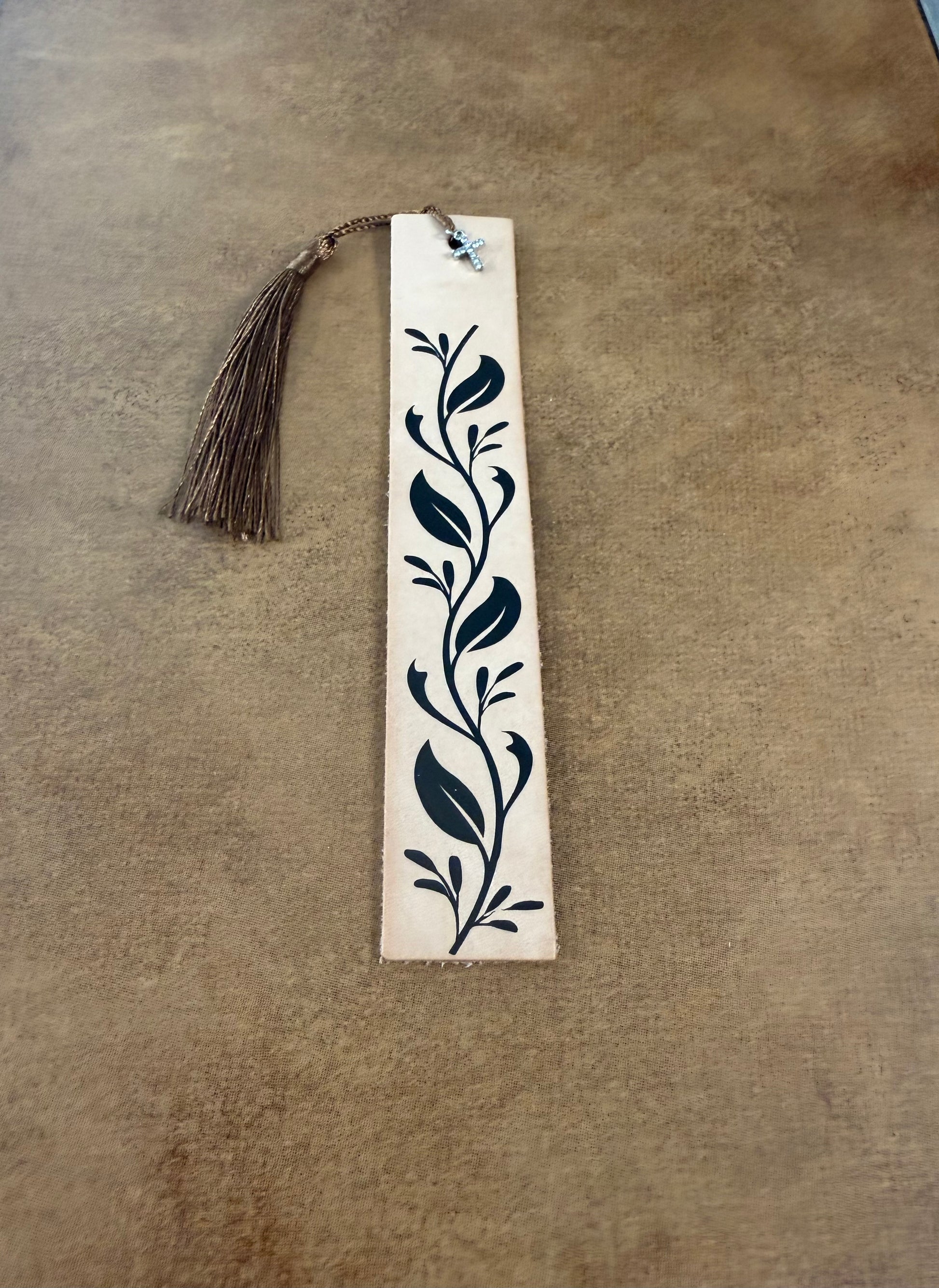 A tan leather bookmark with a black vine pattern, cross charm, and a brown tassel, placed on a neutral background.