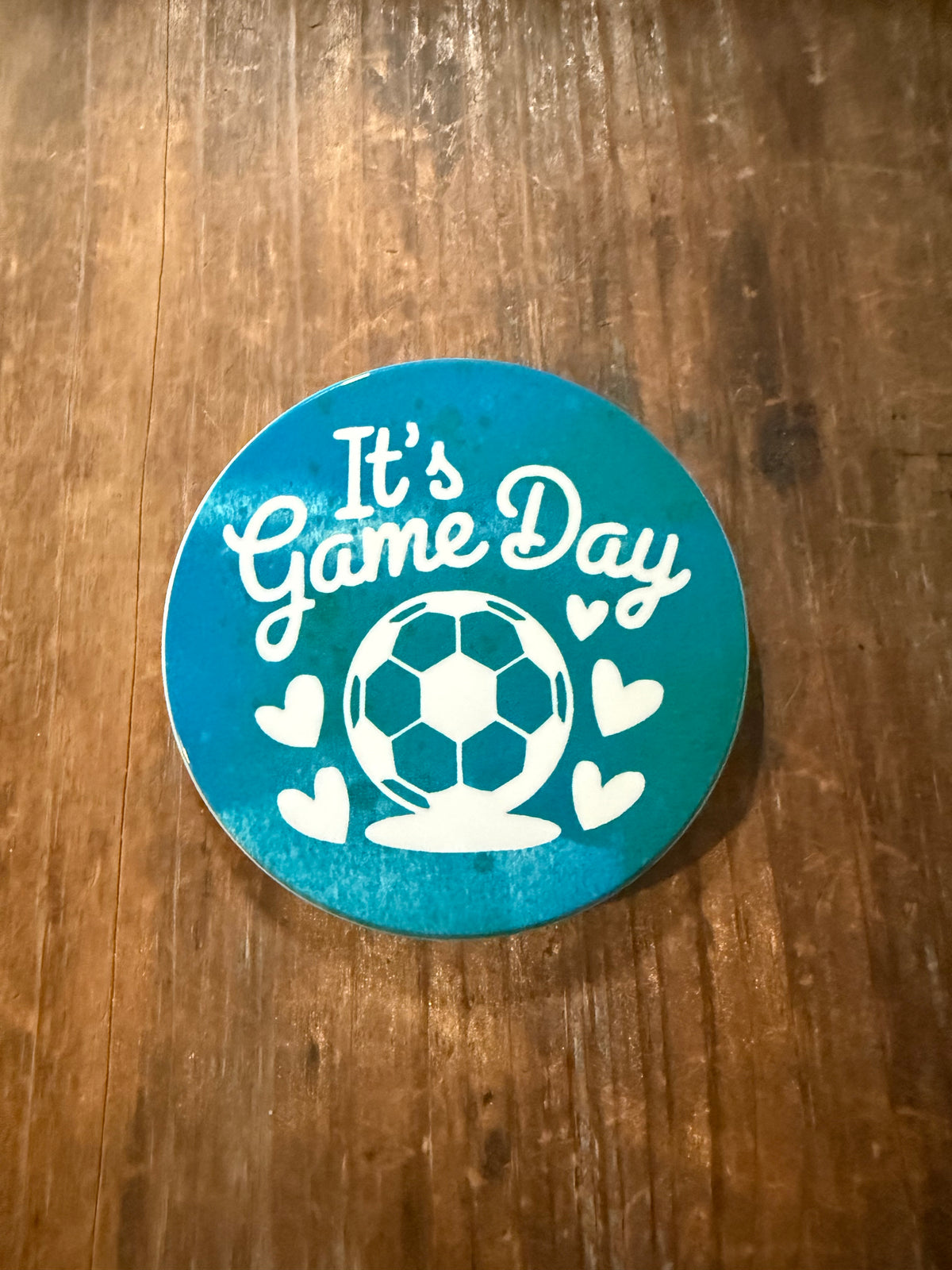 Circular teal coaster with white “It’s Game Day” text, soccer ball graphic, and heart accents on wooden surface. Roxzell
