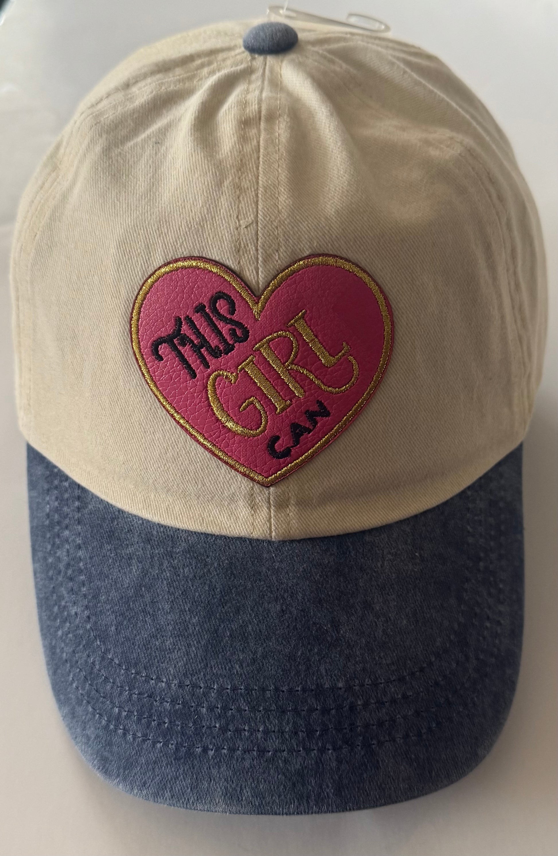 Tan and denim blue all cotton cap with pink and gold heart shaped "This Girl Can" patch on the front. 