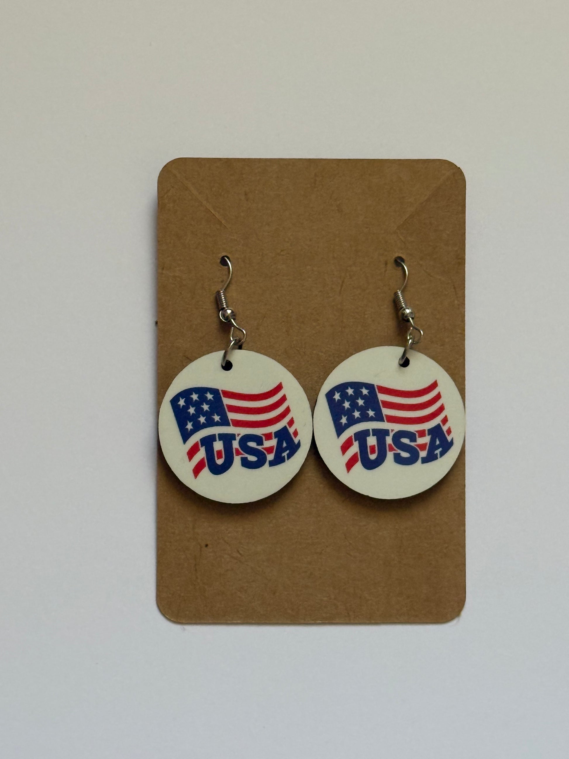 Pair of circular earrings displayed on a brown backing card, each featuring a stylized American flag design with red and white stripes, blue stars, and bold blue “USA” lettering printed across the center.