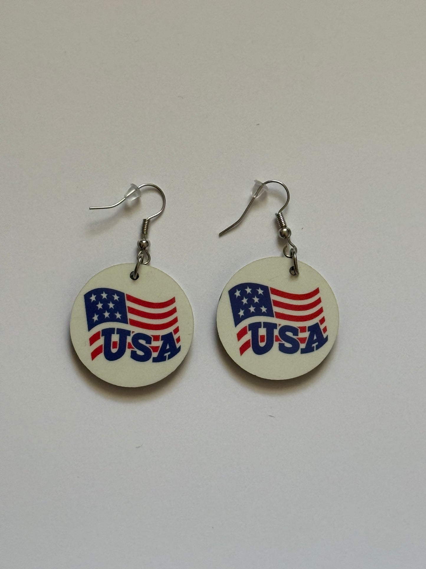 Two round earrings with metal hooks placed on a white background, showing a patriotic design of the American flag and the word “USA” in blue letters on a white surface. Roxzell