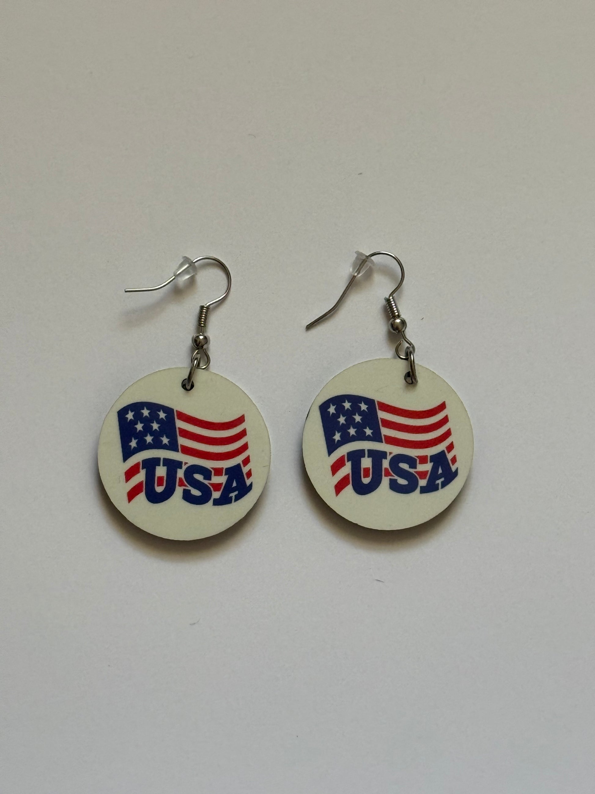 Two round earrings with metal hooks placed on a white background, showing a patriotic design of the American flag and the word “USA” in blue letters on a white surface. Roxzell