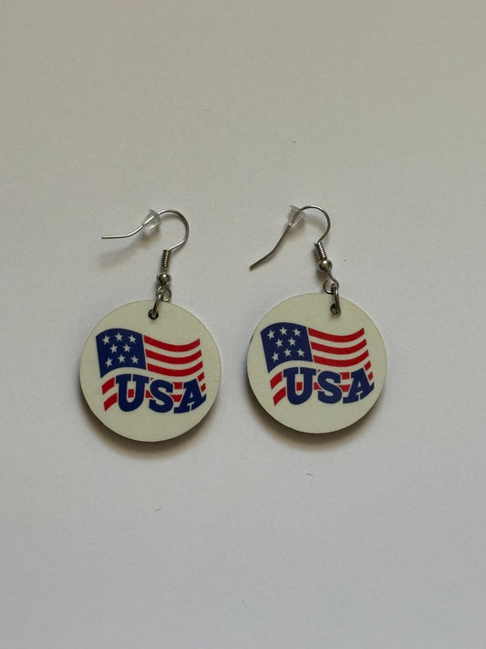 Two round earrings with metal hooks placed on a white background, showing a patriotic design of the American flag and the word “USA” in blue letters on a white surface. Roxzell