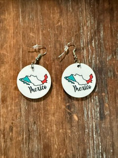 Pair of circular earrings featuring Mexico’s map outline colored in green, white, and red with the word “Mexico” written in cursive, displayed on a wooden surface. Roxzell