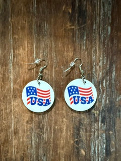 Pair of circular earrings featuring the word “USA!” in bold blue letters with a stylized American flag design of red and white stripes and blue stars, displayed on a wooden surface. Roxzell