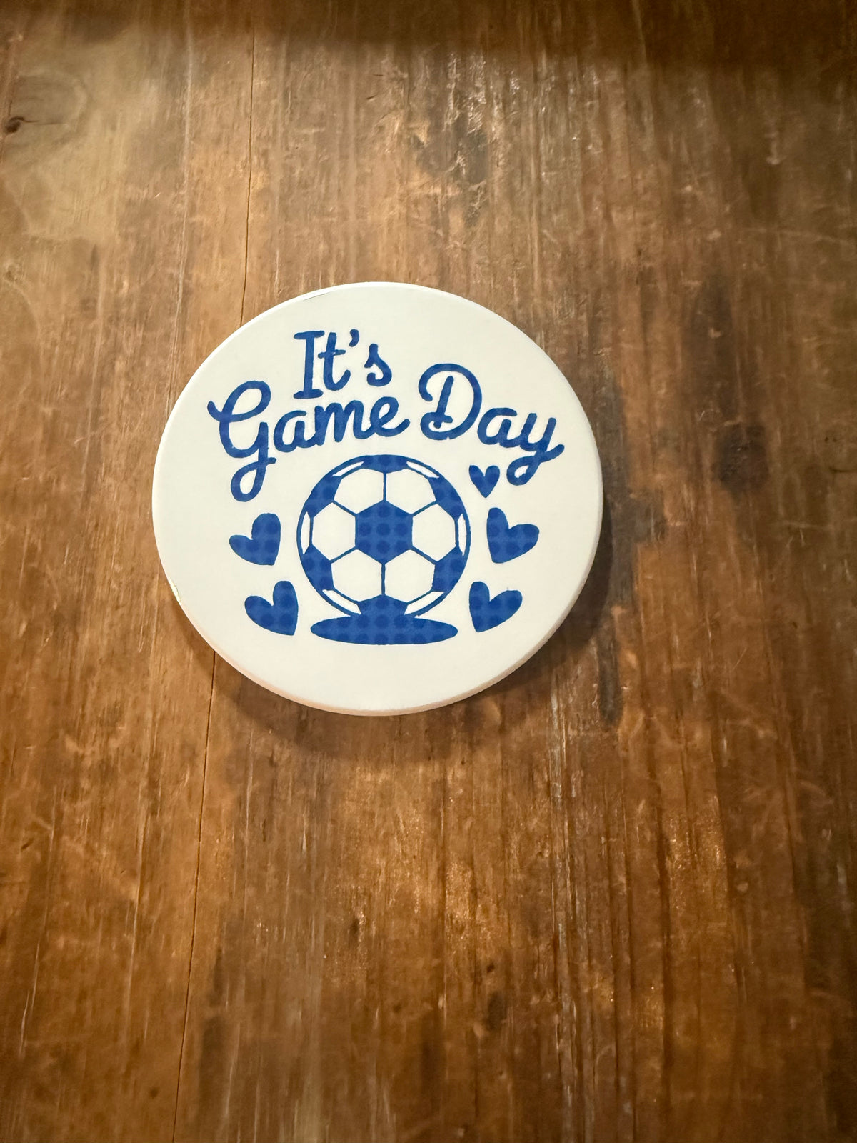 White coaster with blue “It’s Game Day” lettering and soccer ball surrounded by hearts, placed on rustic wood. Roxzell