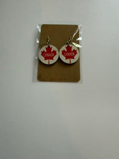 Canada maple leaf earrings attached to a brown backing card inside a clear plastic sleeve, showing red maple leaf designs with “Canada” text across the center.