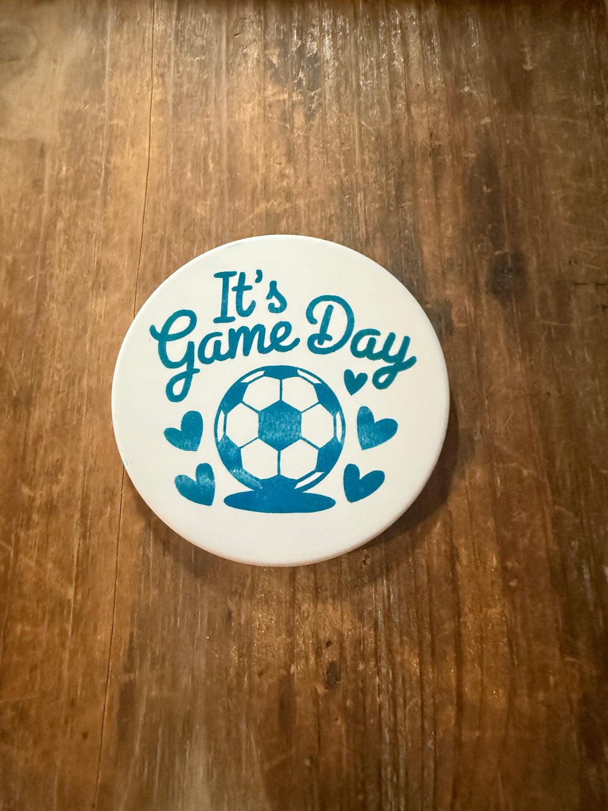White coaster with teal “It’s Game Day” lettering and soccer ball surrounded by hearts, placed on rustic wood. Roxzell