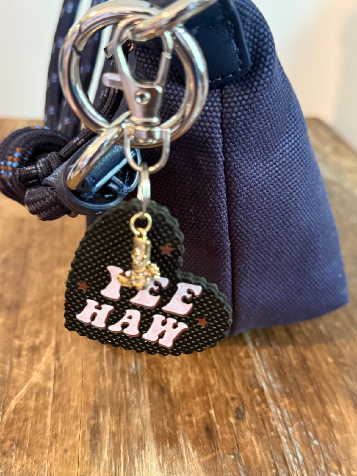 A heart-shaped black keychain charm featuring the words 'YEE HAW' in pink and small brown stars on a navy woven fabric bag. Roxzell