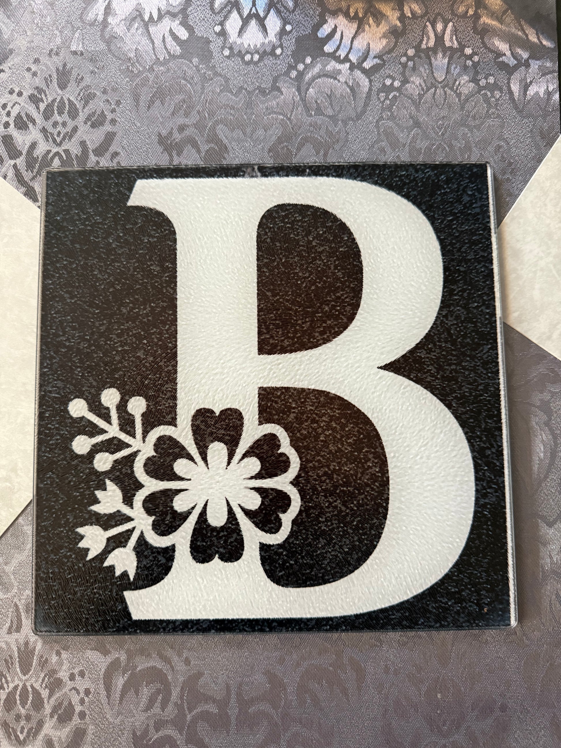 Glass cutting board with uppercase "B" and integrated floral artwork on a gray textured table runner, Roxzell
