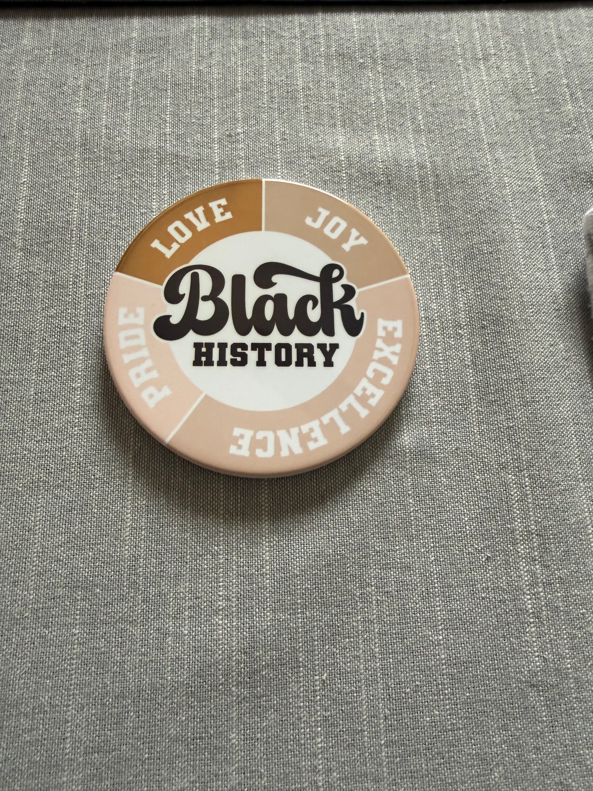Circular Black History coaster with bold Love, Joy, Pride, and Excellence text and colored segments on gray fabric