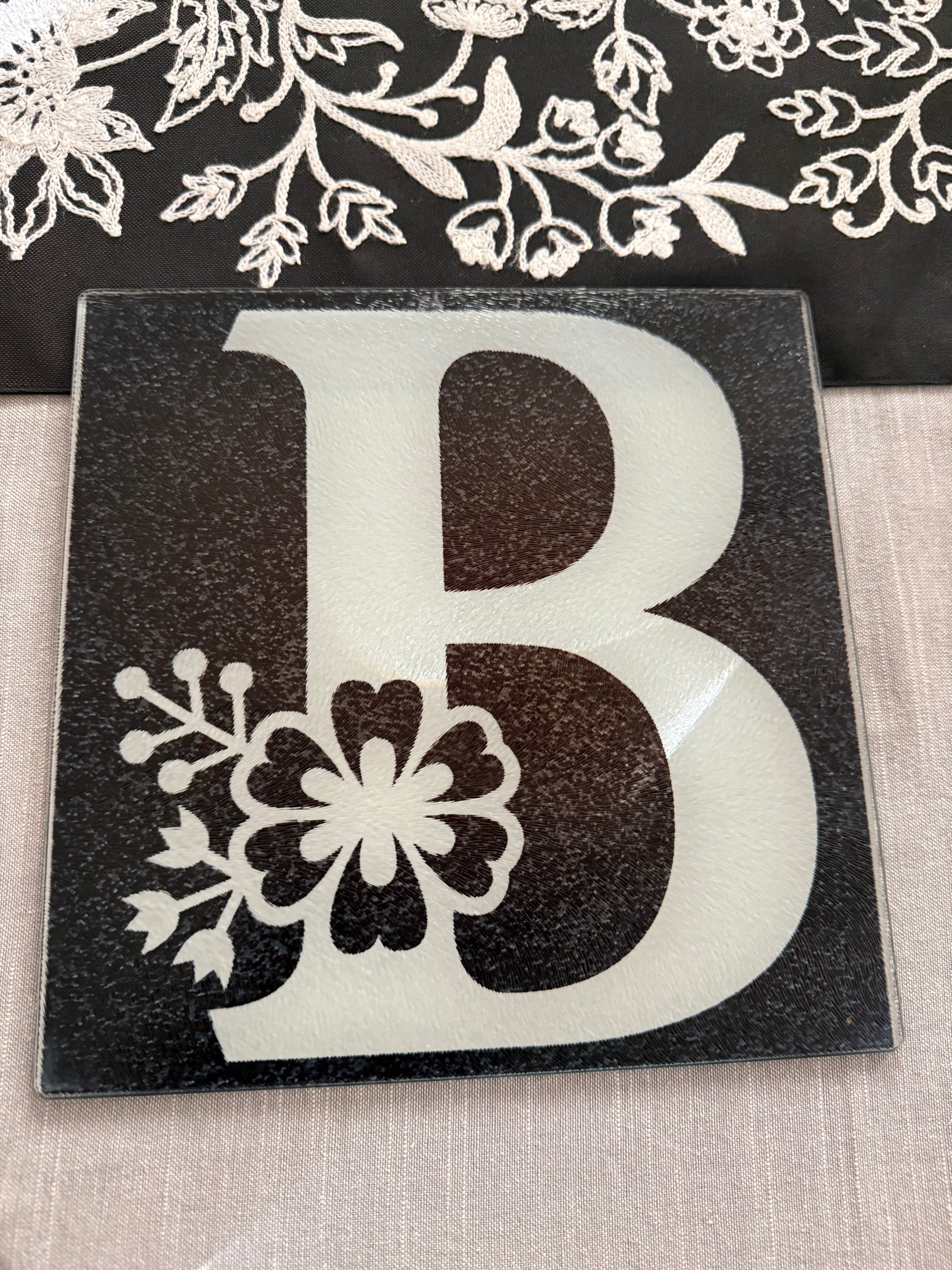 Glass cutting board featuring uppercase "B" with botanical floral design on an embroidered table runner and gray tablecloth, Roxzell
