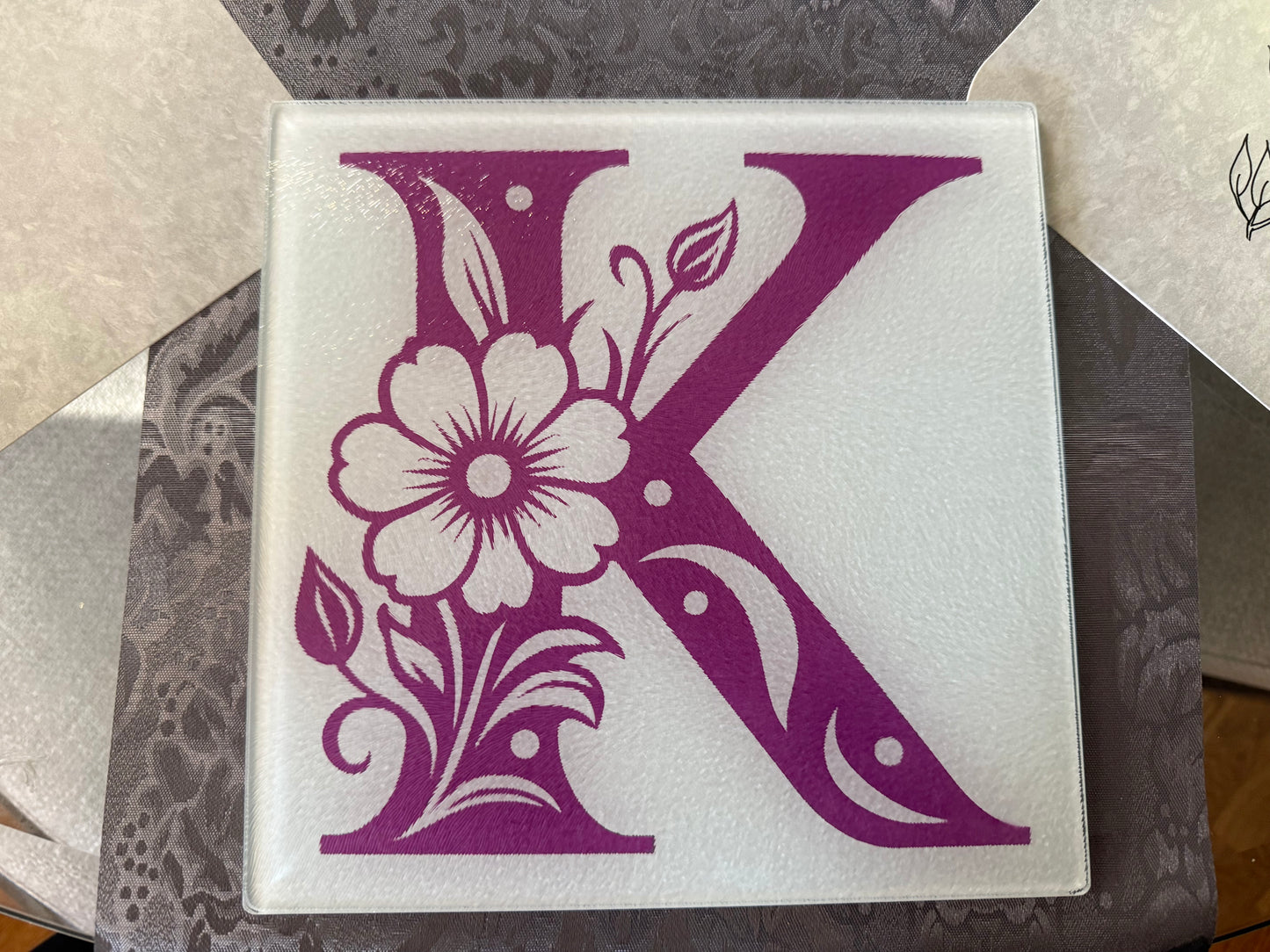 Close-up of decorative purple “K” with flower and leaf details on glass cutting board, Roxzell