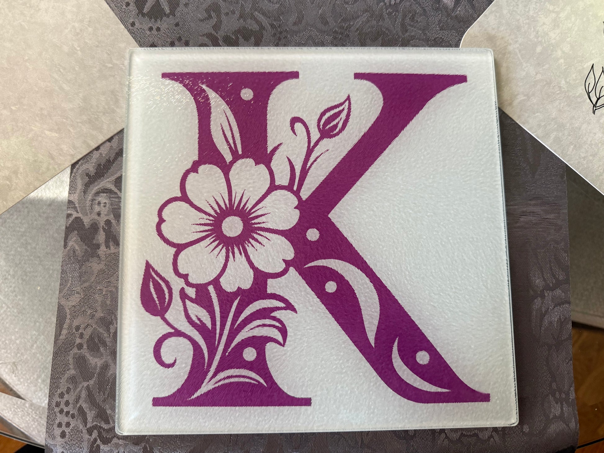 Close-up of decorative purple “K” with flower and leaf details on glass cutting board, Roxzell