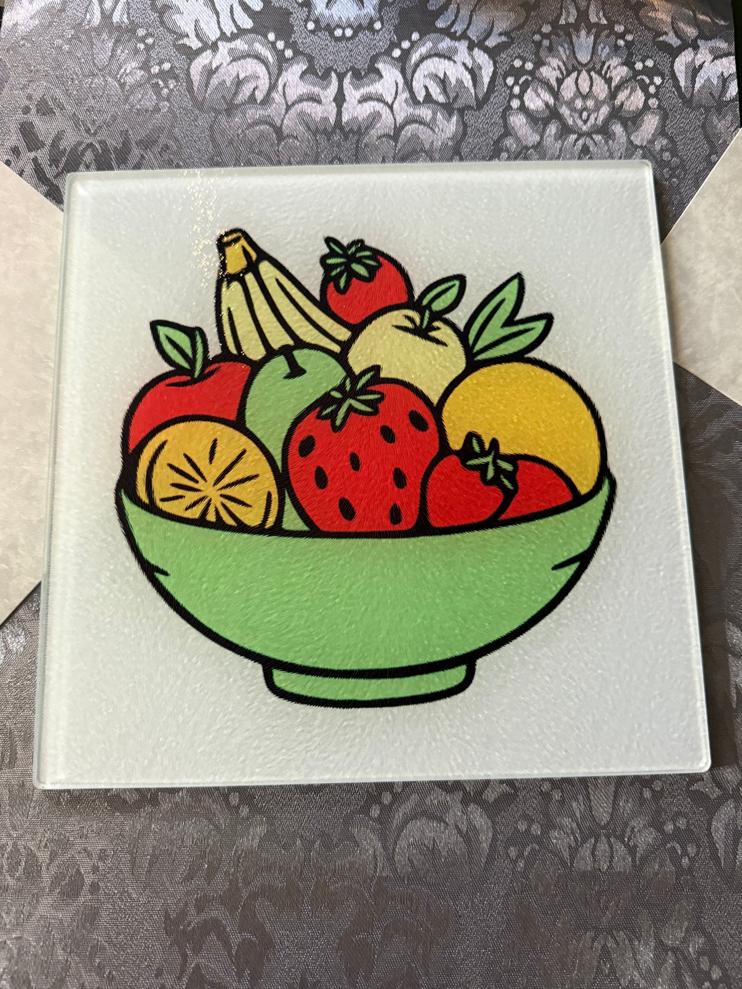 Tempered glass cutting board featuring a vibrant multicolor fruit illustration on a kitchen table with a gray table runner, Roxzell