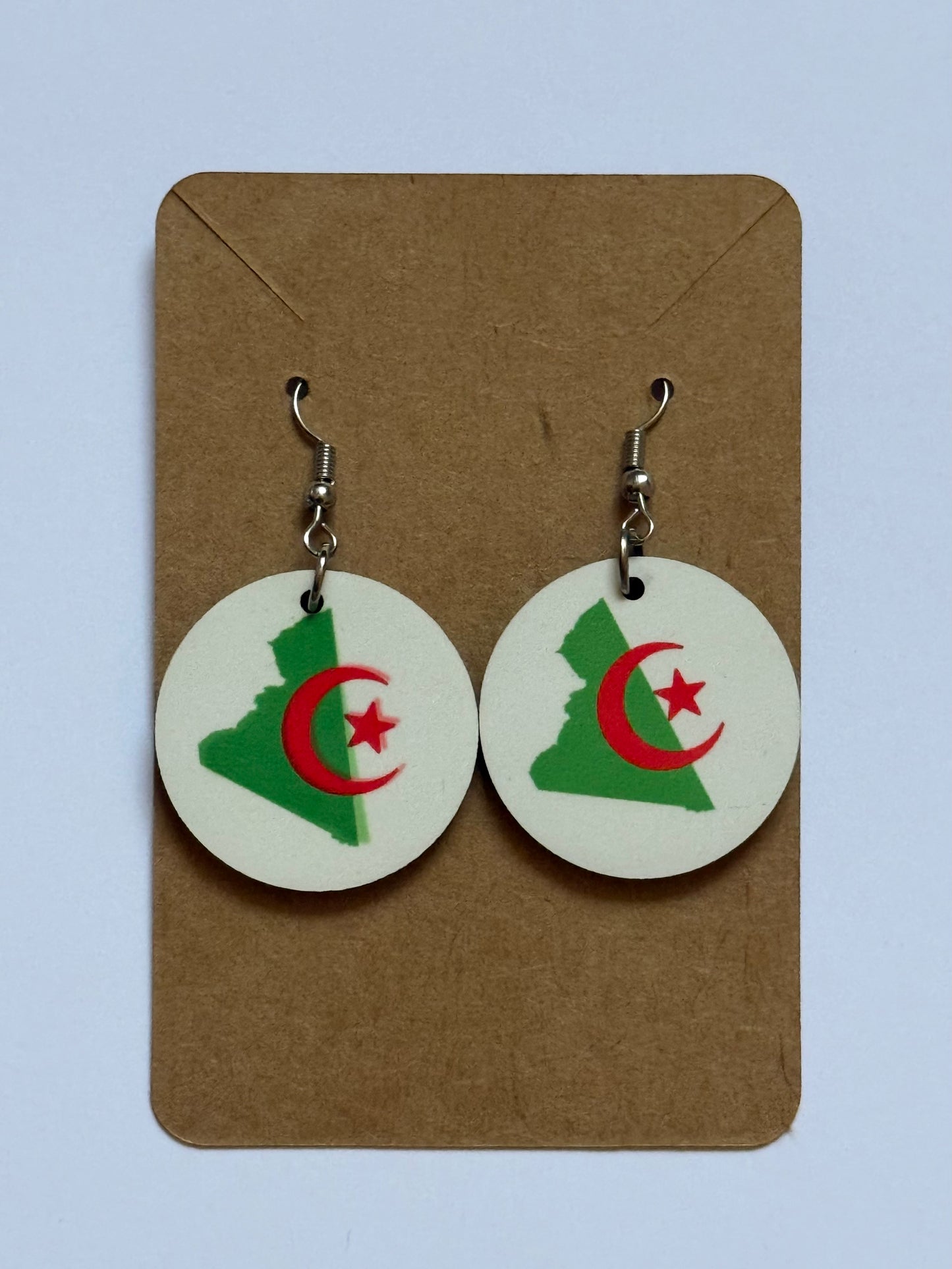 Pair of circular earrings displayed on a brown backing card, each featuring a green silhouette of Algeria’s map with a red crescent and star symbol from the national flag printed on a cream background. Roxzell