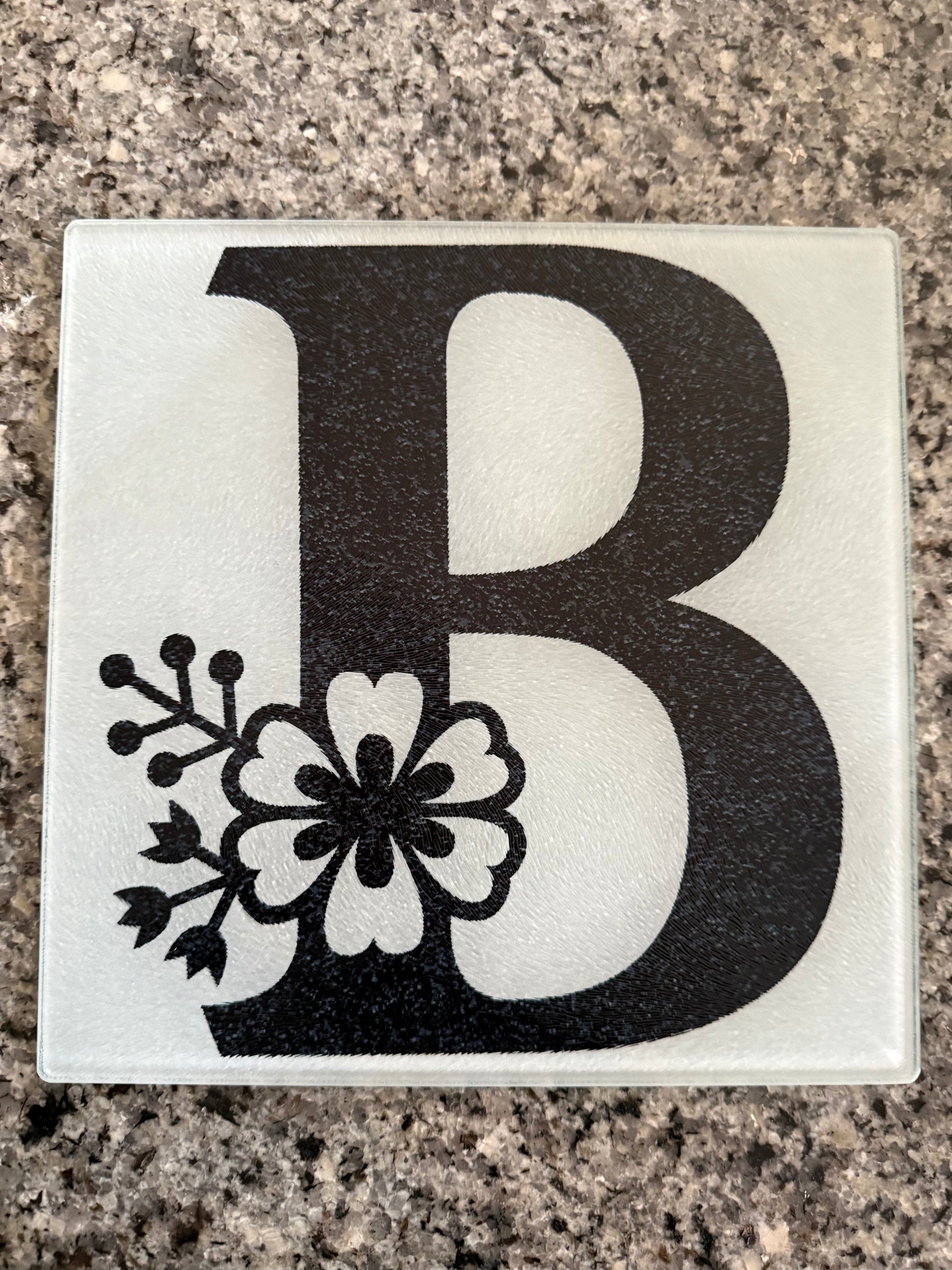 Tempered glass cutting board with black letter "B" and floral embellishment on a granite countertop, Roxzell