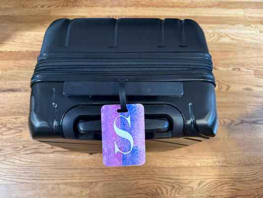 Black suitcase with a purple luggage tag featuring a white 'S' on a wooden floor.