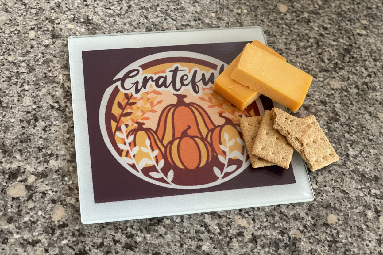 Glass cutting board with pumpkins, fall leaves, and “Grateful” script with cheese and crackers on top of it, Roxzell