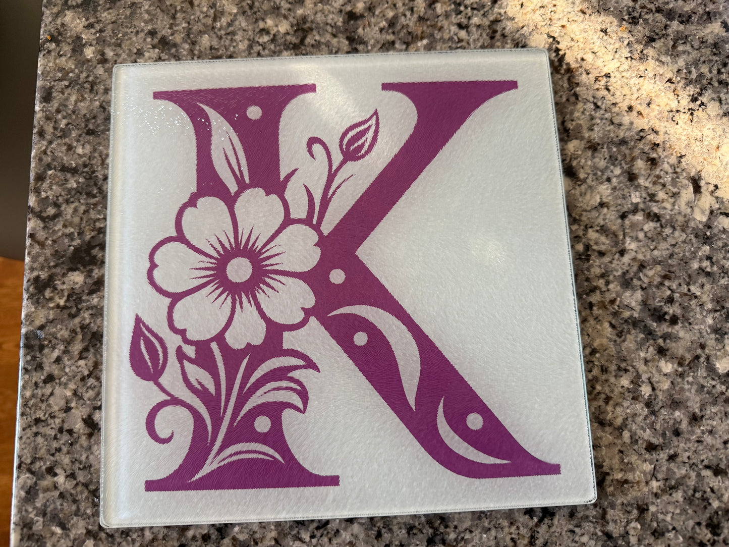 Glass cutting board with purple floral “K” monogram displayed on granite kitchen countertop, Roxzell