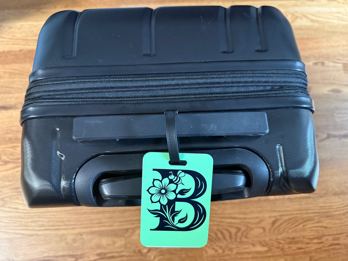 Green luggage tag with floral letter "B" attached to a black suitcase handle, Roxzell
