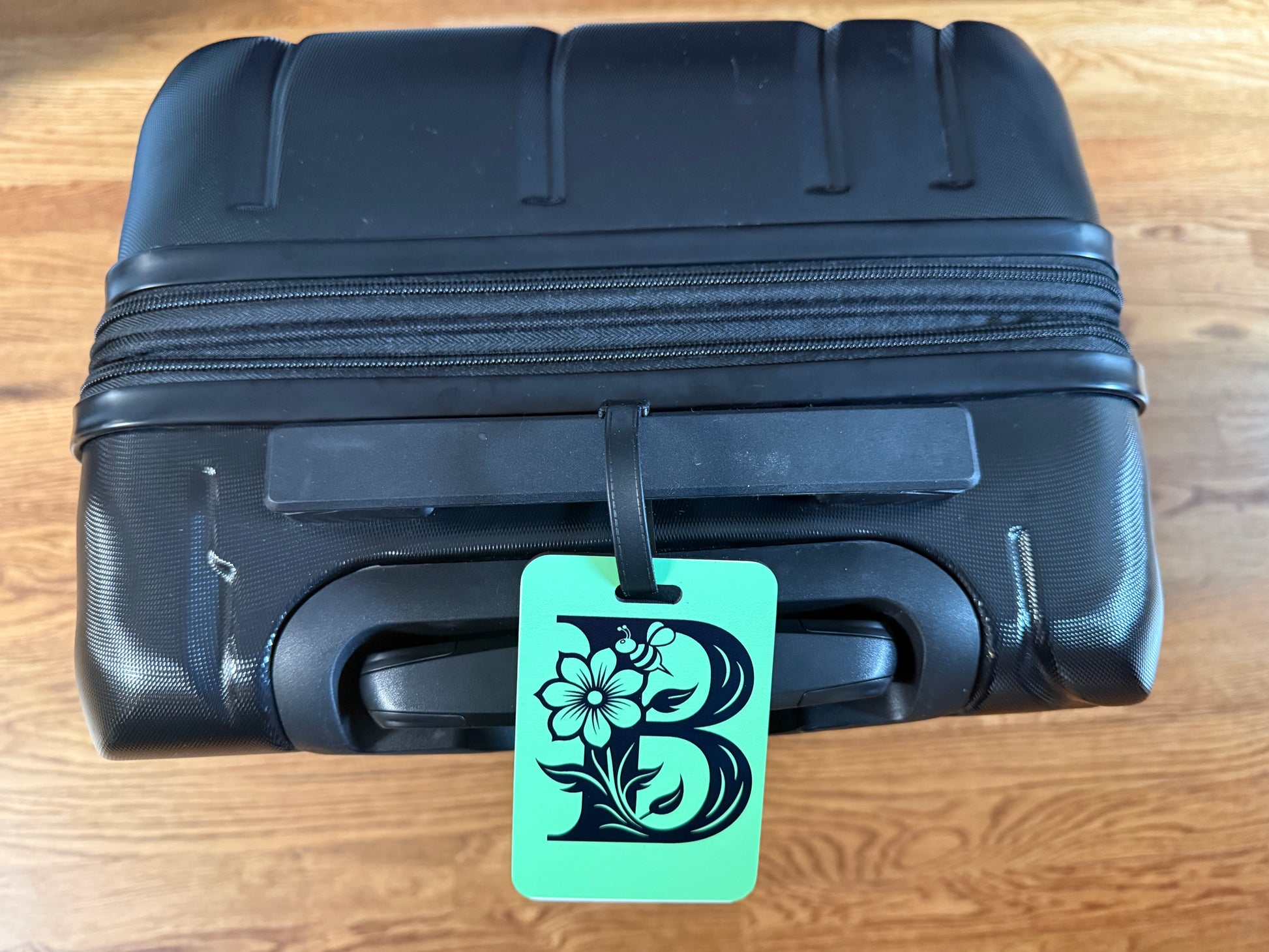 Green luggage tag with floral letter "B" attached to a black suitcase handle, Roxzell
