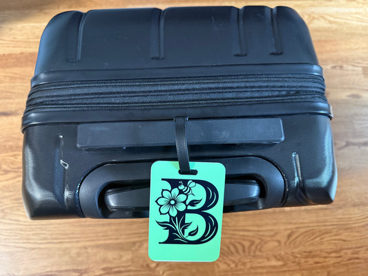 Green luggage tag with floral letter "B" attached to a black suitcase handle, Roxzell
