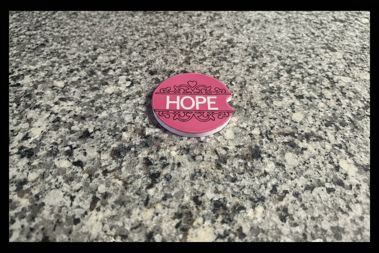 Pink coaster with white “HOPE” lettering and symmetrical heart accents on a granite countertop with a black border. Roxzell