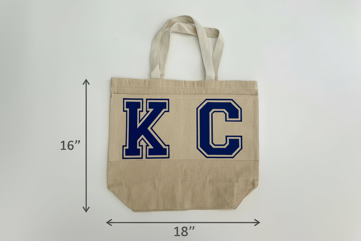 Beige tote bag with blue 'KC' letters and measurements on a light gray background