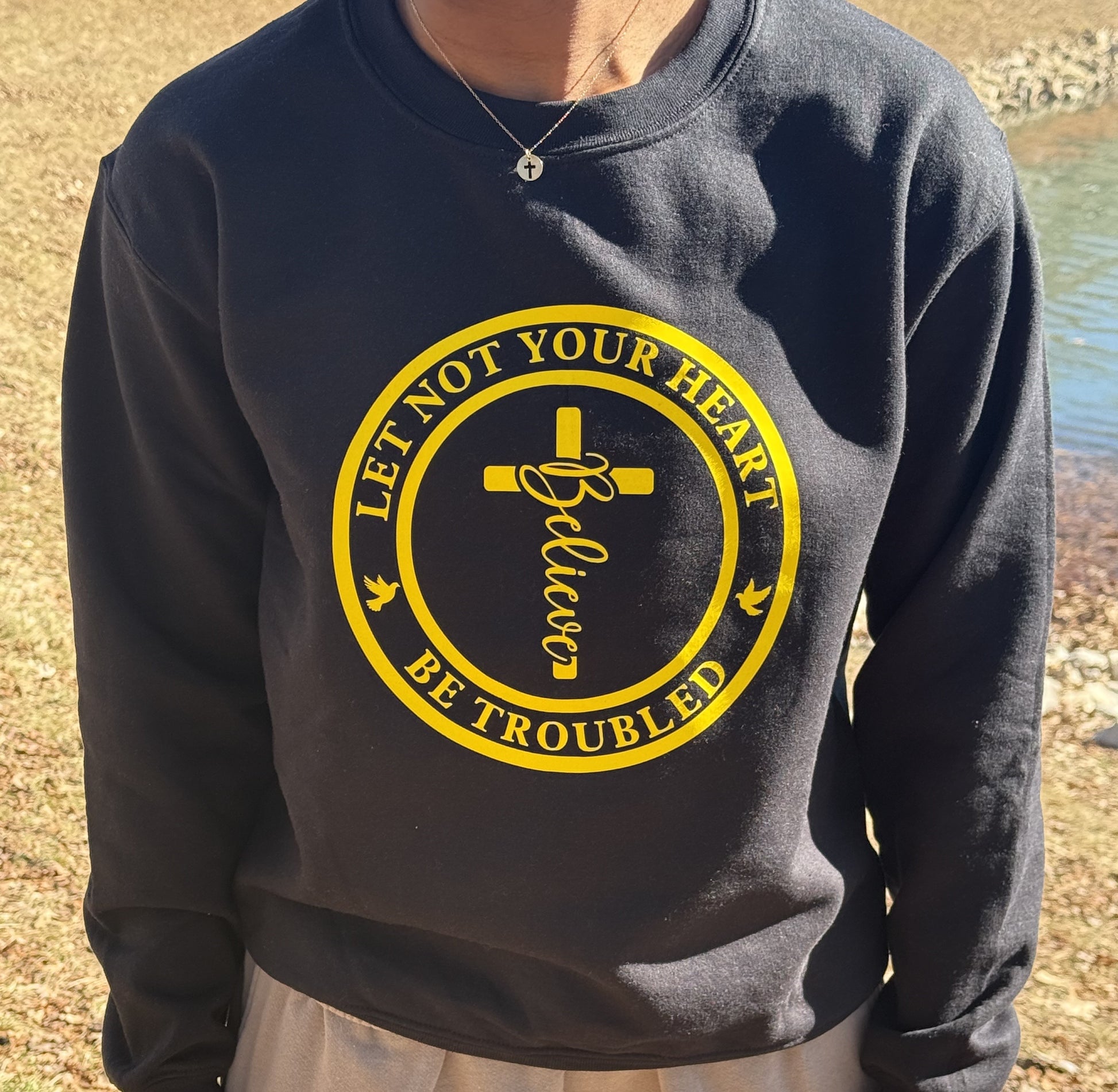 Person wearing a black sweatshirt with yellow cross and “Believe” design, let not your heart be troubled text and doves, Roxzell