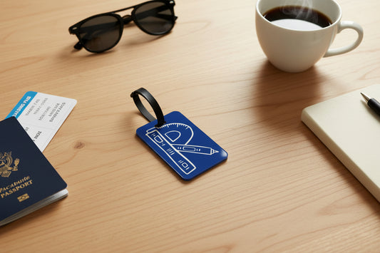 Blue luggage tag featuring white "R" shaped illustrations of a ruler, pen, and protractor on a table with passport and other travel accessories, Roxzell