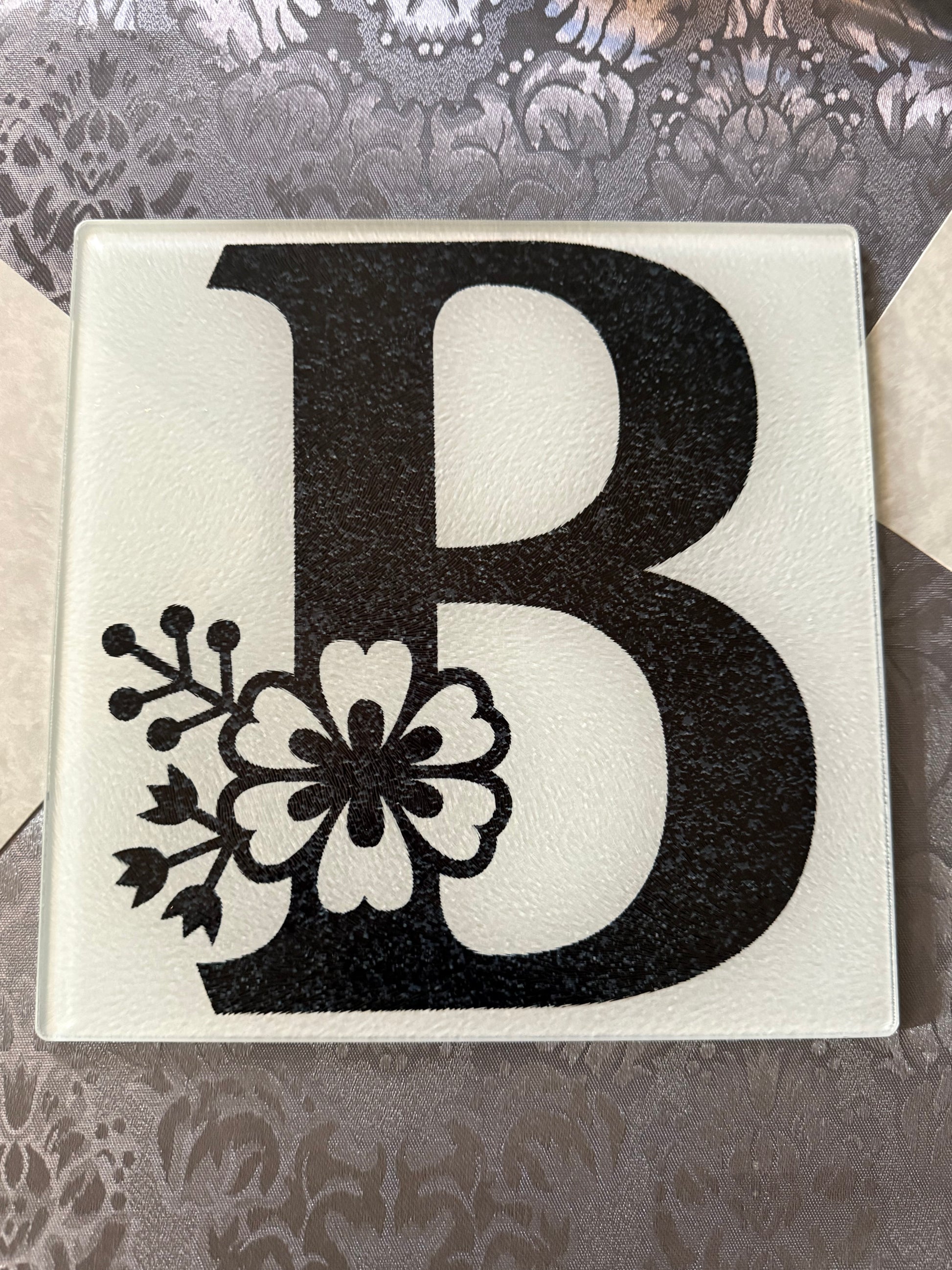 Glass cutting board featuring a black letter "B" with botanical floral design on a gray table runner, Roxzell