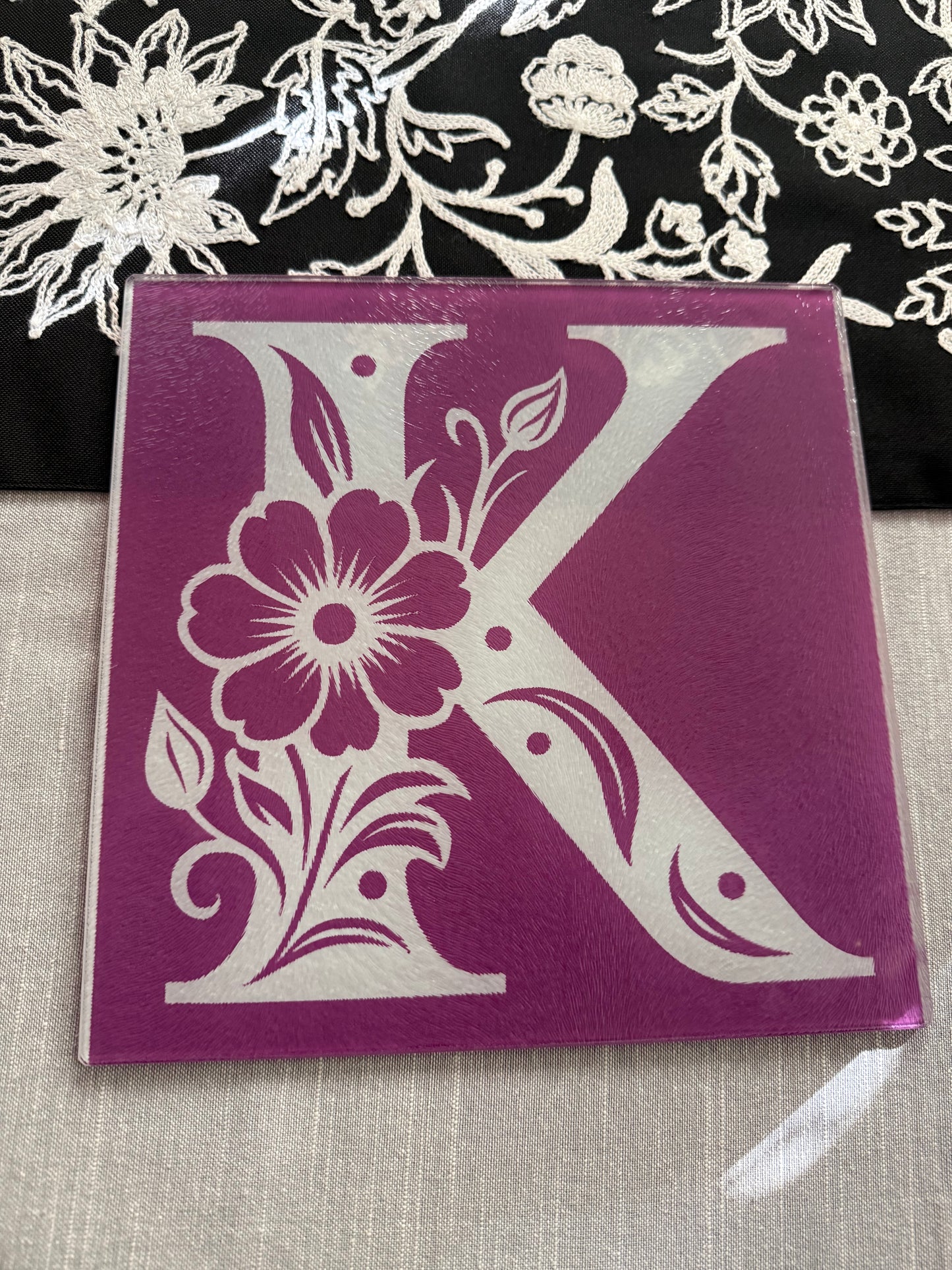 Square glass cutting board with white floral "K" design on purple background, styled with embroidered fabric, Roxzell